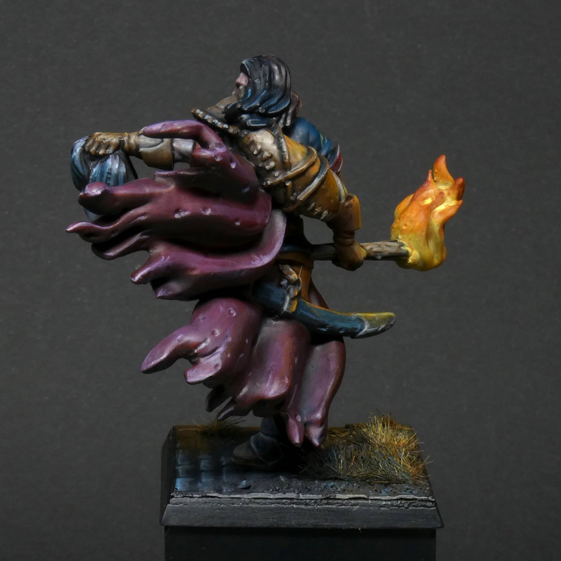 a rear/ back view 32mm scale miniature of Cristof Witchbane, a rugged witch hunter wielding a flaming torch and a severed witch's head.  The model is viewed painted beautifully with excellent lighting effects from the fire of the torch. 